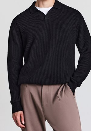 Hombre con jersey negro de punto acanalado con cuello sobre camisa blanca y pantalones beige plisados, mano en el bolsillo.