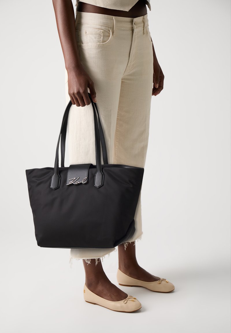 Black tote bag with two short handles, featuring a smooth material, rectangular shape, and a silver logo accent on the front.