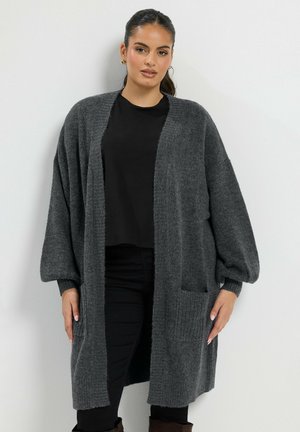 Femme portant un long cardigan gris foncé à manches ballon sur un haut noir et un pantalon noir, debout contre un mur blanc.