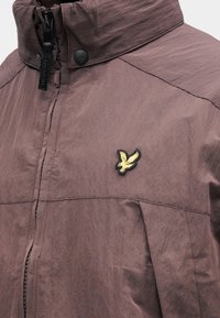 Giacca leggera viola con colletto alto, chiusura con zip e bottoni a pressione. Presenta un logo ricamato di un uccello nero e dorato sul petto.