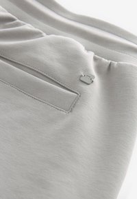 Tissu gris clair avec une poche et un petit logo métallique carré cousu près d'une ceinture élastique sur un vêtement.