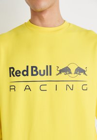 Sudadera amarilla con un audaz logo en gris oscuro de "Red Bull Racing" que presenta dos toros en estilo enérgico, fabricada con una tela suave y un cuello redondo.