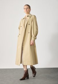 Beige trench coat with a double-breasted design, large buttons, and side pockets, paired with brown heeled ankle boots. Smooth fabric.