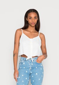Top branco curto com alças finas ajustáveis, featuring um fecho de botão na frente e detalhe de laço na bainha. Combinado com jeans azul claro.