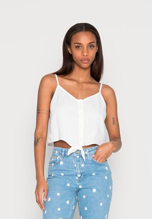 Top branco curto com alças finas ajustáveis, featuring um fecho de botão na frente e detalhe de laço na bainha. Combinado com jeans azul claro.