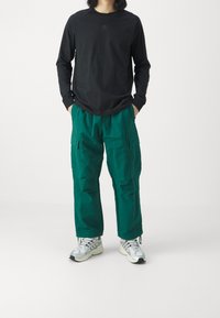 Long-sleeve black shirt with subtle logo, paired with loose-fitting green cargo pants featuring multiple pockets and silver athletic shoes.