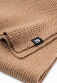Beige ribbet tæppe med en blød tekstur, prydet med et sort mærke med et logo. Stofets overflade udviser et ensartet mønster og varme.