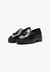 Schwarze Lederloafers mit einer niedrigen Plateausohle, ausgestattet mit einem Riemen über der Oberseite und einem dekorativen Quaste-Detail. Glatte Textur.