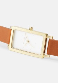 Obdélníkový hodinky ve zlatém tónu s bílým ciferníkem, který má minimalistické hodinové značky. Hnědý kožený pásek s texturovaným povrchem.