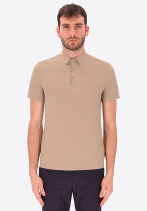 Uomo con capelli corti e scuri che indossa una polo beige a maniche corte e pantaloni scuri, in piedi davanti a uno sfondo chiaro e semplice.