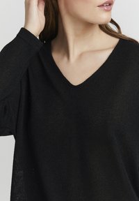 Fekete V nyakú pulóver texturált kötésből, laza szabású és hosszú ujjú. Az anyag könnyűnek és puhanak tűnik.