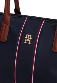 Sac fourre-tout bleu marine avec poignées en cuir marron, rayures verticales rouge-blanc-bleu, et un logo doré entrelacé "TH" près de la fermeture éclair supérieure.