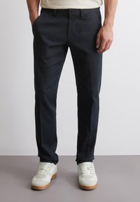 Pantaloni sartoriali blu navy scuro in tessuto liscio, taglio dritto, tasche laterali e una sottile lucentezza, abbinati a sneakers di colore chiaro.
