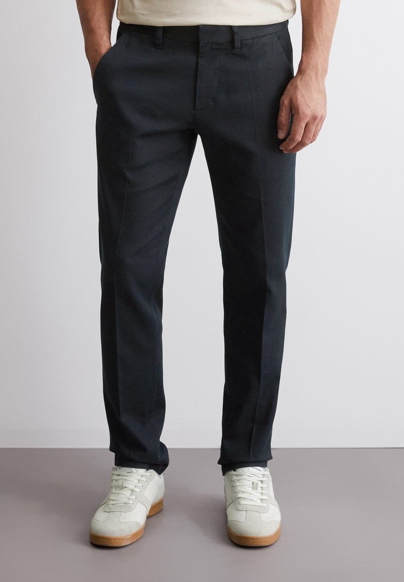 Pantaloni sartoriali blu navy scuro in tessuto liscio, taglio dritto, tasche laterali e una sottile lucentezza, abbinati a sneakers di colore chiaro.