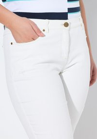 Jeans de mezclilla blanca con detalles desgastados, botones dorados y un diseño ajustado. Los bolsillos son visibles con una mano colocada dentro.