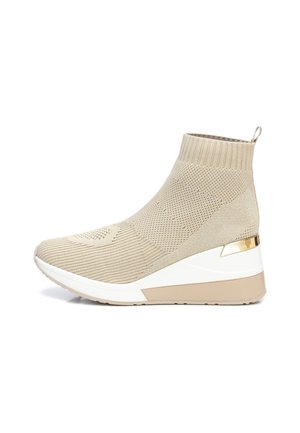 Beigenvärinen neulottu nilkkasaapas valkoisella kiilapohjalla, resorireunuksella, kultaisella yksityiskohdalla kannan lähellä ja vetolenkillä takana.