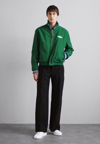 Zelená zipsová bunda s rebrovanými manžetami, nosená cez prúžkovanú košelu. Kombinovaná s čiernymi nohavicami a bielymi teniskami. Jednoduchý dizajn, voľný strih.