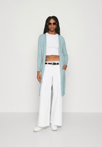 Lyseblå lang cardigan laget av myk strikk, brukt over en hvit cropped topp og parret med hvite vide bukser og hvite sneakers.