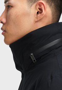 La giacca nera presenta un colletto alto e una tasca con zip. Tessuto liscio, resistente all'acqua, con una finitura opaca e dettagli di cucitura minimi.