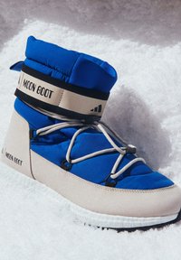 Blå vadderad Moon Boot med en beige bas, som har svarta remmar, grå snören och en texturerad vit sula. Snöbakgrund.