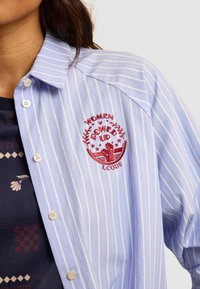 Camicia button-up di un blu chiaro con strisce bianche e rosa. Presenta un ricamo rosso con la grafica "Women Power Up" e bottoni sul davanti.