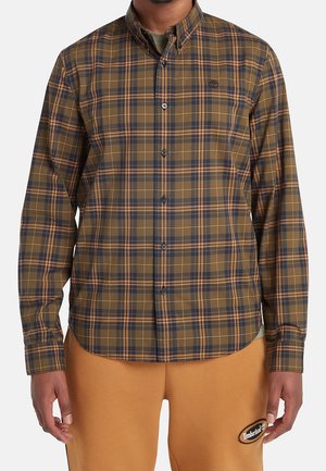 Mężczyzna w brązowej, granatowej i pomarańczowej koszuli w kratę zapinanej na guziki oraz beżowych spodniach z naszywką z logo Timberland na lewym udzie.