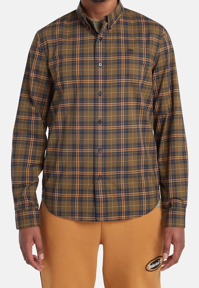 Homme portant une chemise à carreaux marron, bleu marine et orange à boutons avec un pantalon beige présentant un patch logo Timberland sur la cuisse gauche.