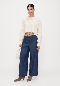 Woman wearing a cream cable-knit cropped sweater, wide-leg blue pants with drawstring, gray ankle boots, gold hoop earrings, and carrying a small white bag.
