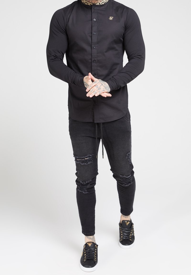 Homme portant une chemise noire à manches longues boutonnée avec un col à motifs, un jean noir déchiré et des baskets noires à semelles blanches.