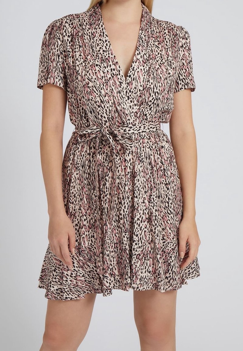 Femme portant une robe à manches courtes, col en V, ceinturée, avec un motif léopard rose et noir, debout devant un fond uni clair.