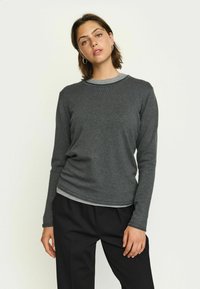 Maglione grigio a maniche lunghe con texture a coste e scollo rotondo, indossato sopra una camicia grigia più chiara, abbinato a pantaloni neri.