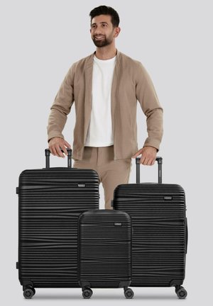 Un homme en veste et pantalons beiges se tient avec trois valises rigides noires de différentes tailles, à roulettes, contre un fond gris.