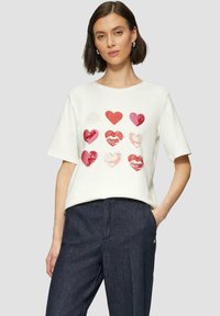 Witte katoenen T-shirt met verschillende harten in rode en roze tinten, korte mouwen en een ronde halslijn. Gepaard met donkerdenim broek.