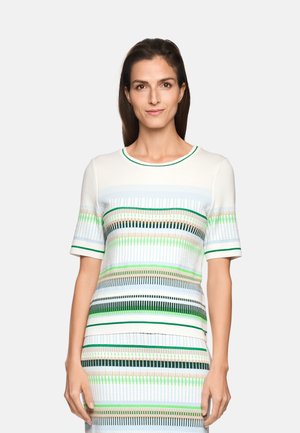 Frau mit glattem braunem Haar, die ein weißes Kurzarm-Top und einen Rock mit horizontalen Streifen in Grün, Blau, Beige und Schwarz trägt.