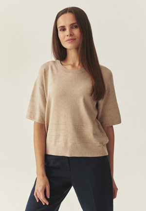 Kvinde med langt, glat brunt hår iført en beige kortærmet striktrøje og mørke bukser, stående mod en ensfarvet lys baggrund.