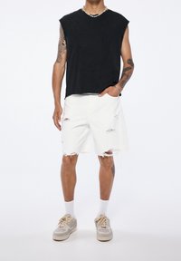 Top de algodón negro sin mangas, pantalones cortos blancos desgastados con bordes deshilachados y zapatillas grises con acentos beige. Fondo blanco liso.