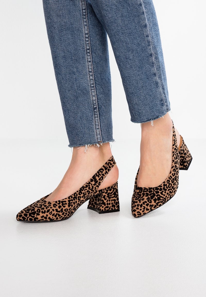 Pieds portant un jean bleu raccourci et des chaussures slingback à talon bloc imprimé léopard sur un sol blanc.
