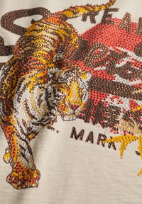 Tijgerafbeelding versierd met meerkleurige strasssteentjes op beige stof, met een gedeeltelijk rood en geel cirkelvormig ontwerp en zwarte tekst.