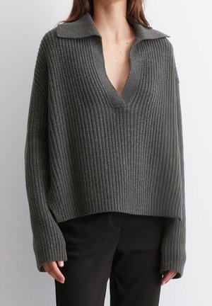 Kobieta ubrana w luźny, ciemnoszary prążkowany sweter z szerokim kołnierzem i głębokim dekoltem w serek, w połączeniu z czarnymi spodniami na jednolitym tle.