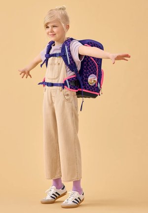 Junges Mädchen mit blondem Haar trägt beige Latzhosen, ein lila Shirt und Turnschuhe, streckt die Arme weit aus und hat einen blau-pinken Rucksack dabei.