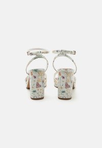 Sandalias blancas de tacón bloque con tiras en el tobillo con un colorido diseño floral y de mariposas, mostradas desde atrás sobre un fondo blanco.