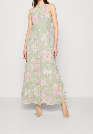 Kvinde iført en ærmeløs pastelgrøn maxikjole med pink blomsterprint og blondebånd i taljen, kombineret med matchende grønne højhælede sandaler.