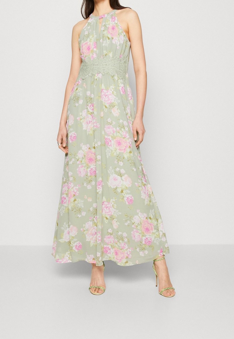 Femme portant une robe maxi sans manches vert pastel avec imprimé floral rose et ceinture en dentelle, assortie à des sandales vertes à talons hauts.