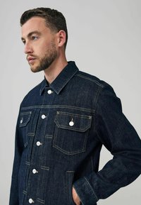 Tumman denim-takkia, jossa on napitettava etuosa, kaksi rintataskua ja pistemallinen kaulus. Erottuva keltainen tikkaus ja teksturoitu kangas.