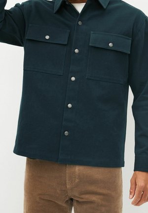 Chemise en velours côtelé vert foncé avec deux poches sur la poitrine et des bouton-pression en argent, dotée d'un col classique et d'une surface texturée.