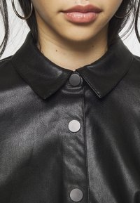 Czarna skórzana koszula z ostrym kołnierzem, matowymi guzikami i gładką teksturą. Widok z bliska pokazuje szczegóły kołnierza i guzików.