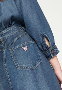 Mid-blue farkku mekko, jossa on tiukka vyötärö, pitkät hihat ja avoimet hihansuut. Takataskussa on punavalkoinen kolmiomainen logo-patch.