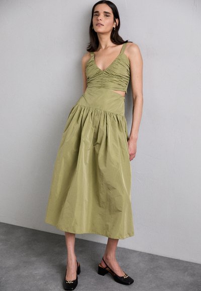 Vestido verde oliva con cuerpo fruncido, escote en V profundo, cintura entallada y falda fluida. Presenta elegantes tacones negros con un acento dorado.