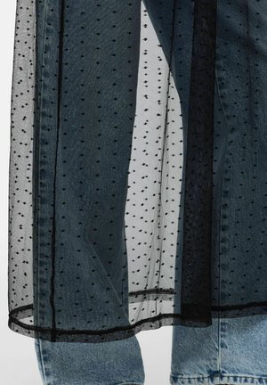 Tessuto nero trasparente a pois sovrapposto a jeans di denim azzurro chiaro, che mostra il contrasto di texture e il dettaglio dell’abbigliamento a strati.