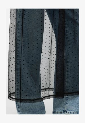 Tessuto nero trasparente a pois sovrapposto a jeans di denim azzurro chiaro, che mostra il contrasto di texture e il dettaglio dell’abbigliamento a strati.